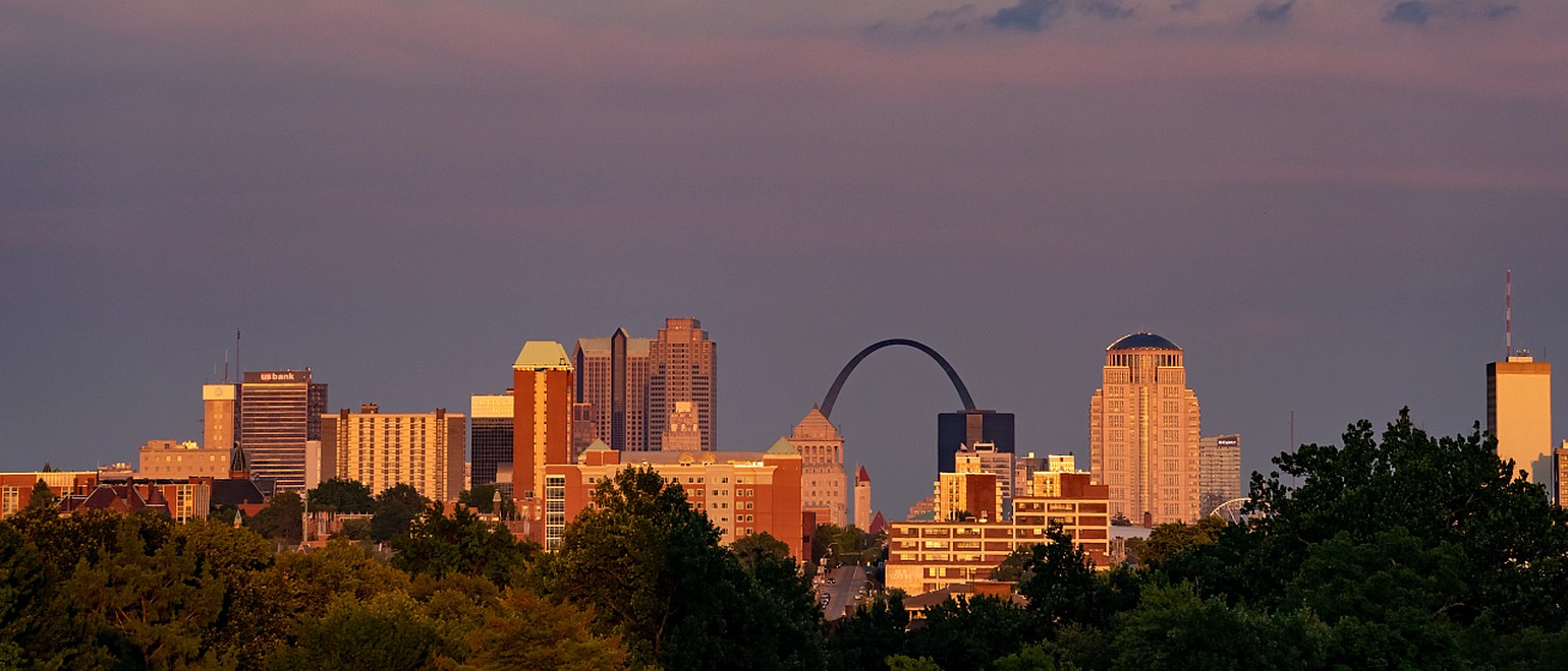 The downtown St. Louis skyline at sunset, with the Gateway Arch rising behind the office towers and a wooded ridge in the foreground